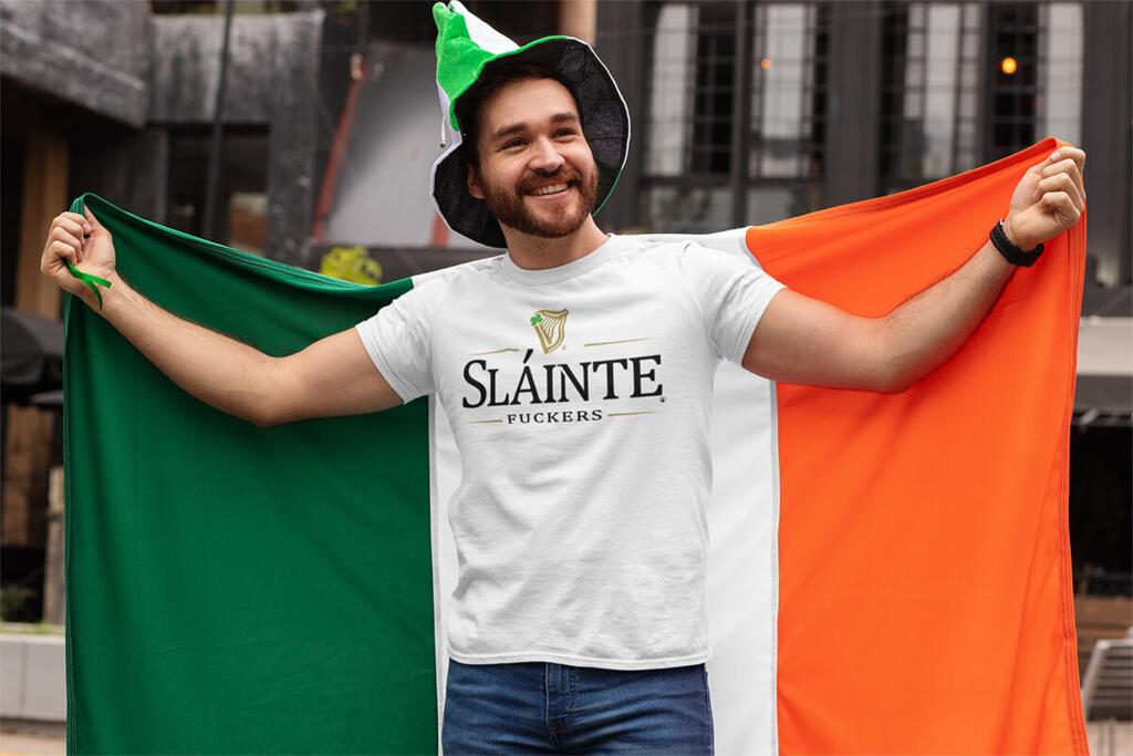 Young man holding irish flag and wearing a white tshirt with SLAINTE FUCKERS design by © 2021 funnydrinking.com